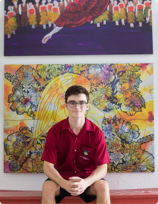 A young man in a red shirt sits on a bench, with paintings on the wall behind him