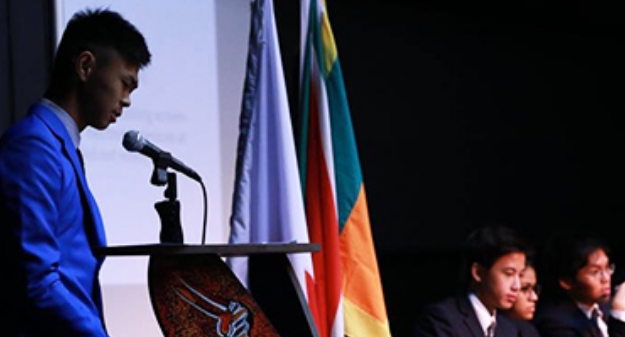 A suited AISM student speaks at a podium, flanked by others, during the World Scholars Cup, highlighting co-curricular engagement.