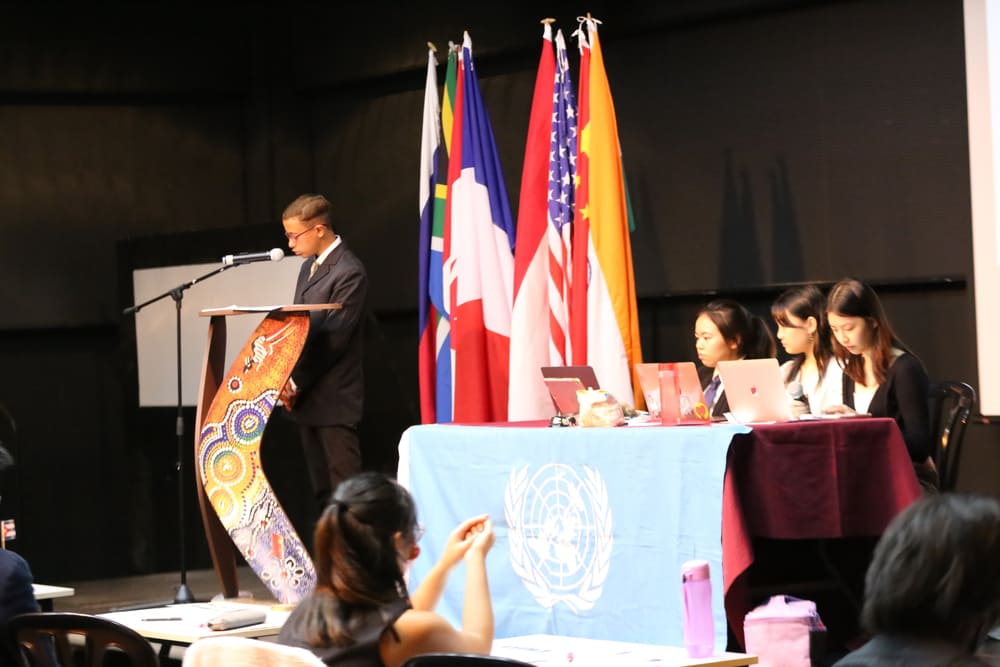 One of the co-curricular activities in school: Students deliver s a speech in front of a podium while attendees sit at tables, engaged and attentive to the presentation.
