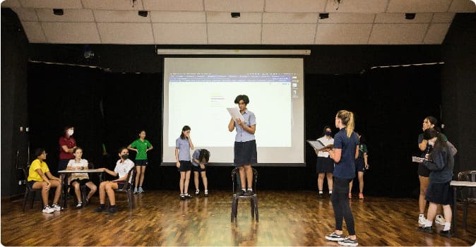 AISM performing arts class with students on stools in front of a screen, engaging in co-curricular activities.